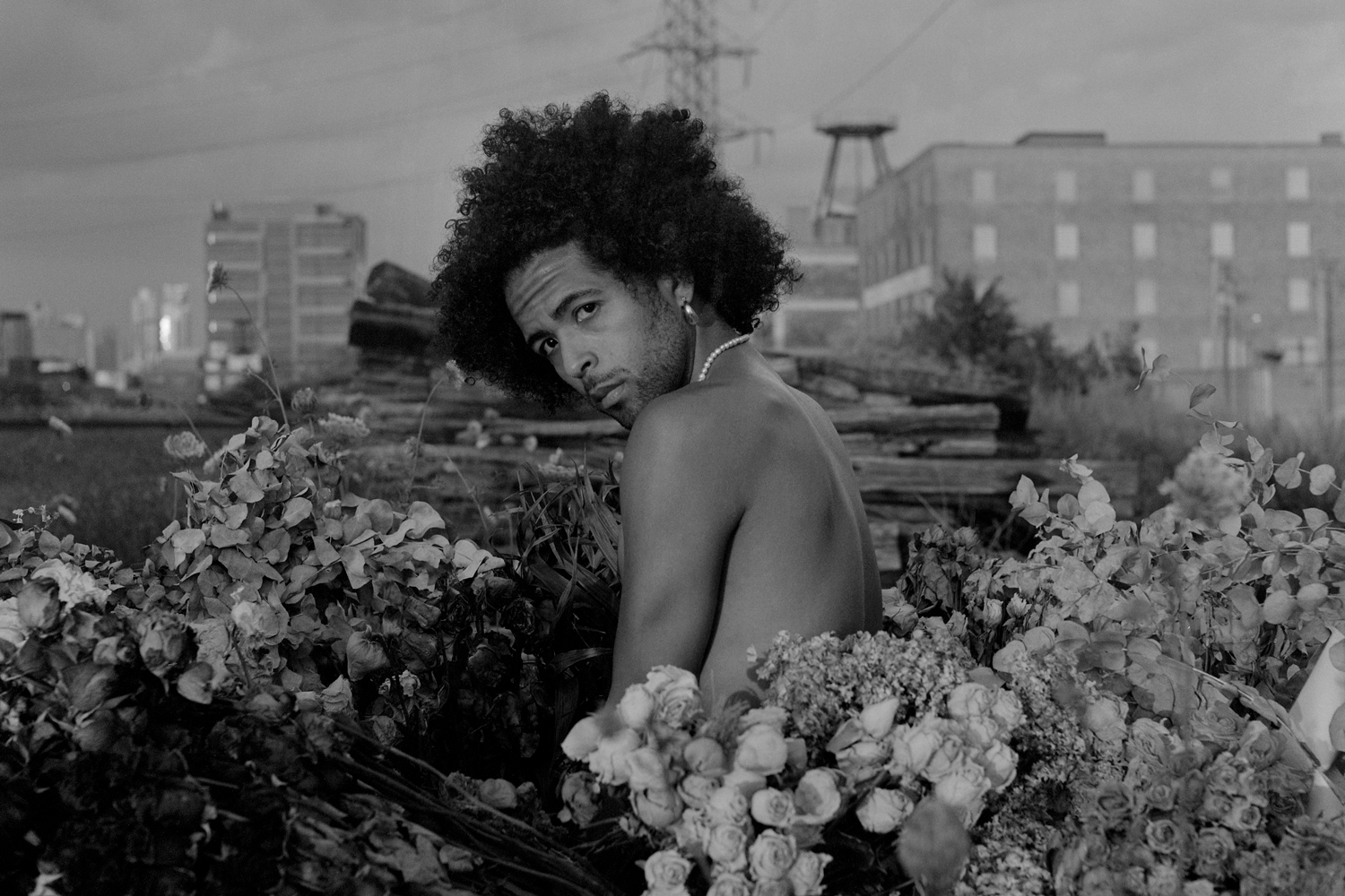 Image: A black-and-white portrait of Erol looking directly into the camera and sitting among dry flowers outdoors on an elevated train track. Buildings and a cloudy sky can be seen in the background. Photo by Ryan Edmund Thiel.
