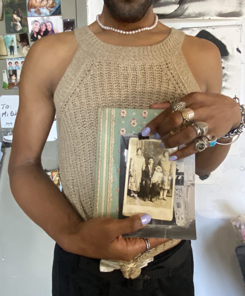 Image: A snpashot in Erol's studio during the interview. Erol is seen from the mouth down, holding his aunt's journals and family photos in his hands. In the background there are more family photos, notes, and drawings taped up on a door. Photo by Tempestt Hazel.