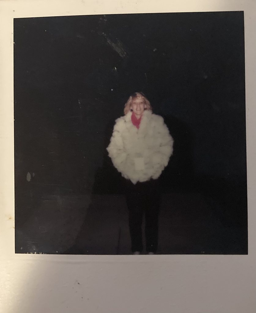 Image: A polaroid image of a young blonde woman clad in a white fluffy coat. She stares at the viewer with a smile. Image courtesy of Gabrielle Lynch.