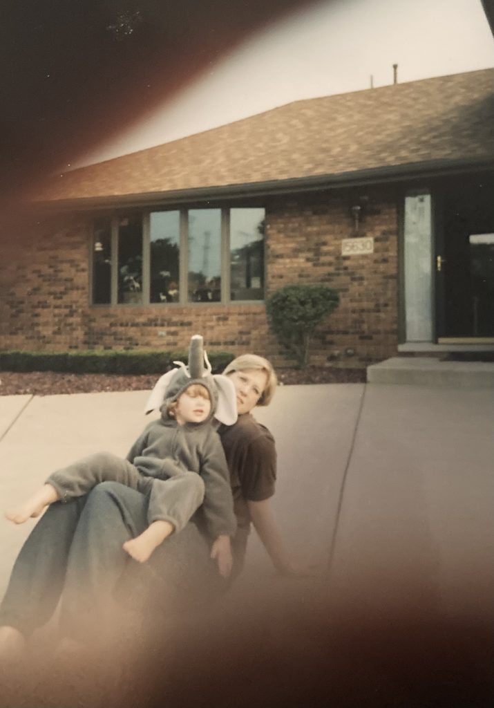 Image: A young woman sitting in the driveway in front of a brown brick house. In her lap is a small child dressed as an elephant. Image courtesy of Gabrielle Lynch.