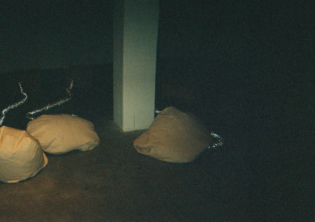 Image: A film photo of Maya Dunietz’s installation, Mechanical Lungs (2022). Three brown bags with metallic silver chords protruding from the tops lie in the center left. Two of the bags are closer together, separated from their third by a white column in the center of the photo. On the other side of the white column is the third bag. The third bag has the least amount of silver chord visible, while the other two have silver chords extending past the white column. All three bags are installed on the gallery floor. Photo by the author.