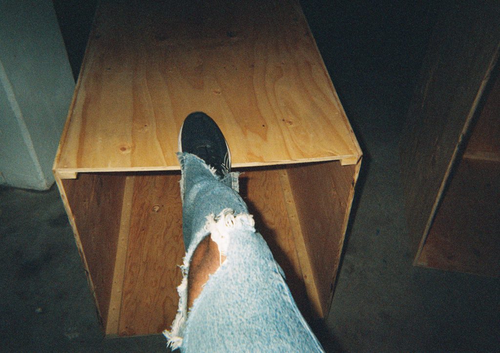 Image: A film photograph of Maya Dunietz’s piece, Boxes, located in gallery four of Bemis Center for Contemporary Arts. The photo shows two plywood boxes. One box is cut out of the frame on the right side. The other box is in the main foreground. Jared Brown’s right leg is photographed planted on the top of the box. Brown is wearing light blue acid wash jeans with a big hole in the knee and the side edges of the jeans cut, with black and white tennis shoes. Photo by the author.