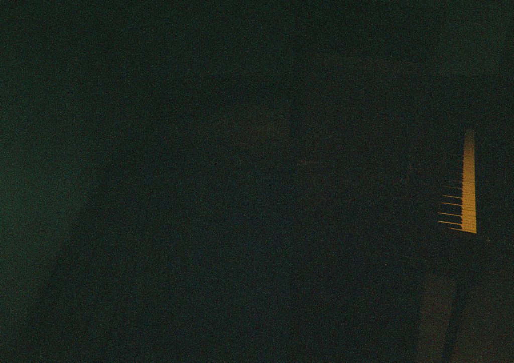 Image: A film photograph of Maya Dunietz’s Root of Two installation at Bemis Center for Contemporary Arts. A grainy, dark photograph shows a piano on the right side of the photo. The most legible part of the piano are the keys. Very faintly on the left, there are chords on the ground. Photo by the author.