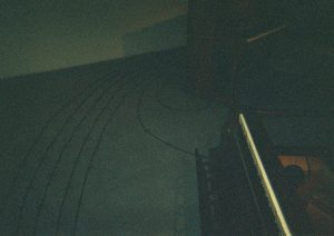 Image: A film photograph of Maya Dunietz’s Root of 2 installation at Bemis Center for Contemporary Arts. A grainy, dark photograph shows two pianos on the far right side of the composition. At the bottom right lies a piano on its back. The piano keys are facing upwards, and the black keys blend in with the rest of the piano’s dark features. The other piano is oriented conventionally above. There are no visible characteristics of the piano sitting upright. To the left of both pianos are seven black chords on the ground. Five of the chords create a track or musical bar from the bottom left of the image, extending to the top right of the photo. There are two chords that appear to be connected to both pianos in the photograph. Photo by the author.