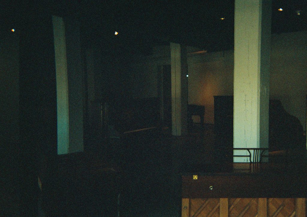 Image: A film photograph of Maya Dunietz’s Root of 2 installation at Bemis Center for Contemporary Arts. A dark, grainy photograph shows various pianos oriented in different ways. The bottom right of the photo shows the back of a brown (wooden) piano: lighter wood with diagonal wood paneling and a darker brown wood at the top with a stand attached where sheet music would rest. The rest of the photo has dark silhouettes of various pianos. There are three white columns in the photograph, almost forming a triangle. Light hits the white panel on the furthest right of the photo. Photo by the author.