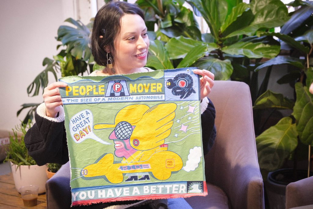 Image: Julia Arredondo holding up an artwork by Atlan Arceo-Witzl, during filming of QTVC Live! The piece that is being held up is on fabric and has a person saying, "HAVE A GREAT DAY!" on it. The text "PEOPLE MOVER" and "YOU HAVE A BETTER" can also be read on the piece. Julia looks to the right side of the composition while smiling. Photograph by Jet Vong.