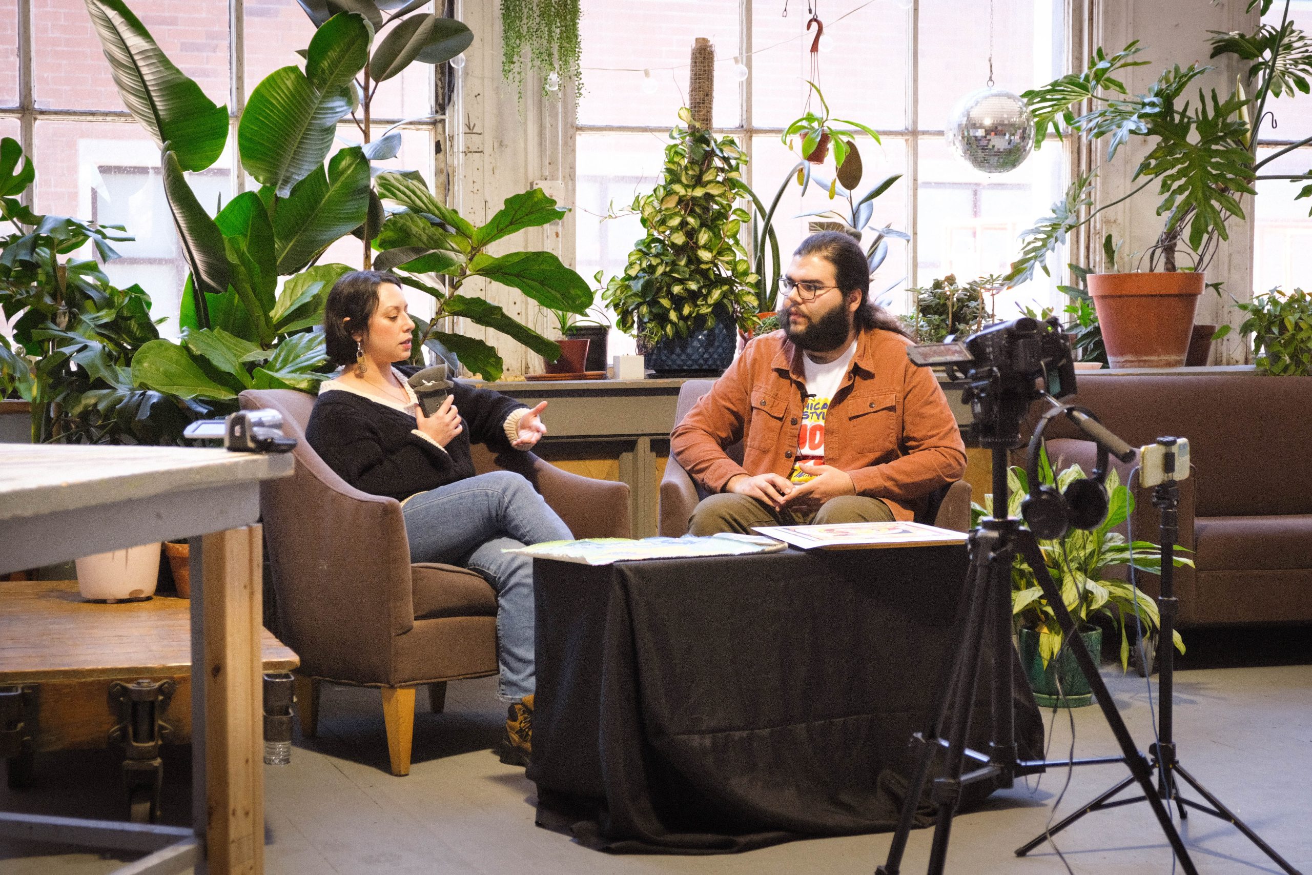 Image: Julia Arredondo and Atlan Arceo-Witzl on the set of QTVC Live! The two face each other while talking and sitting on brownish chairs. Various plants sit behind them while cameras sit in front of them. Photograph by Jet Vong.