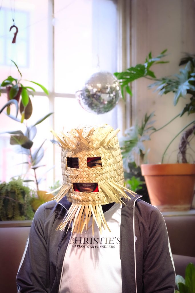 Image: CHema Skandal on the set of QTVC Live! CHema wears a face mask made of straw and a shirt that says, "CHRISTIES exploits art handlers". Photograph by Jet Vong. 
