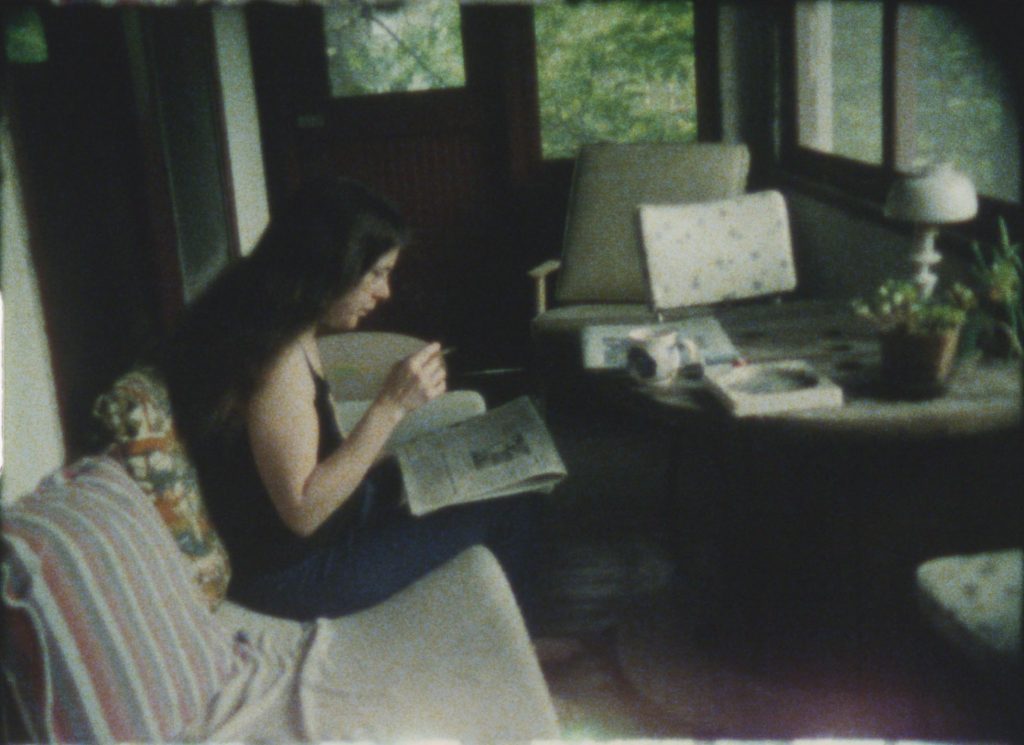 Image: Still image from Melon Patches, or Reasons to Go On Living. A young woman is seated on a couch in a small sitting room. On her lap is a newspaper that she reads intently. Courtesy of the Anne Charlotte Robertson Collection, Harvard Film Archive, Harvard University.