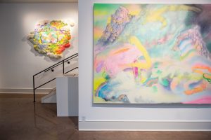 Image: Installation view of Rachel Collier's Still Together. A large, colorful, abstract painting hangs in the foreground, and a round, organic-shaped, colorful piece hangs in the background to the left. Courtesy of Saint Kate Arts Hotel.