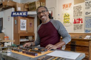 Image: Ben Blount smiles at the camera while standing at his printing press in his studio. Behind him are prints of his hanging on the wall. Photo by Ryan Edmund Thiel.