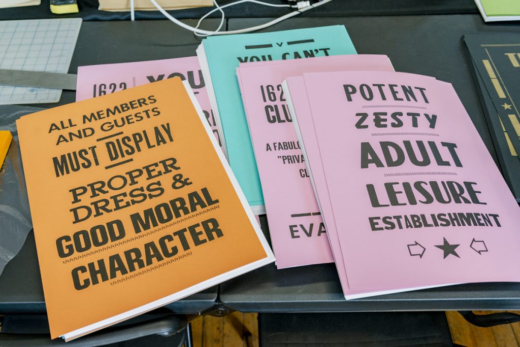 Image: Large flyers in various colors (soft pink, orange, light teal) sit on a table. An orange poster on the left reads: "All members and guests must display proper dress & good moral character." A soft pink poster on the right reads: "Potent, zesty adult leisure establishment." Photo by Ryan Edmund Thiel.