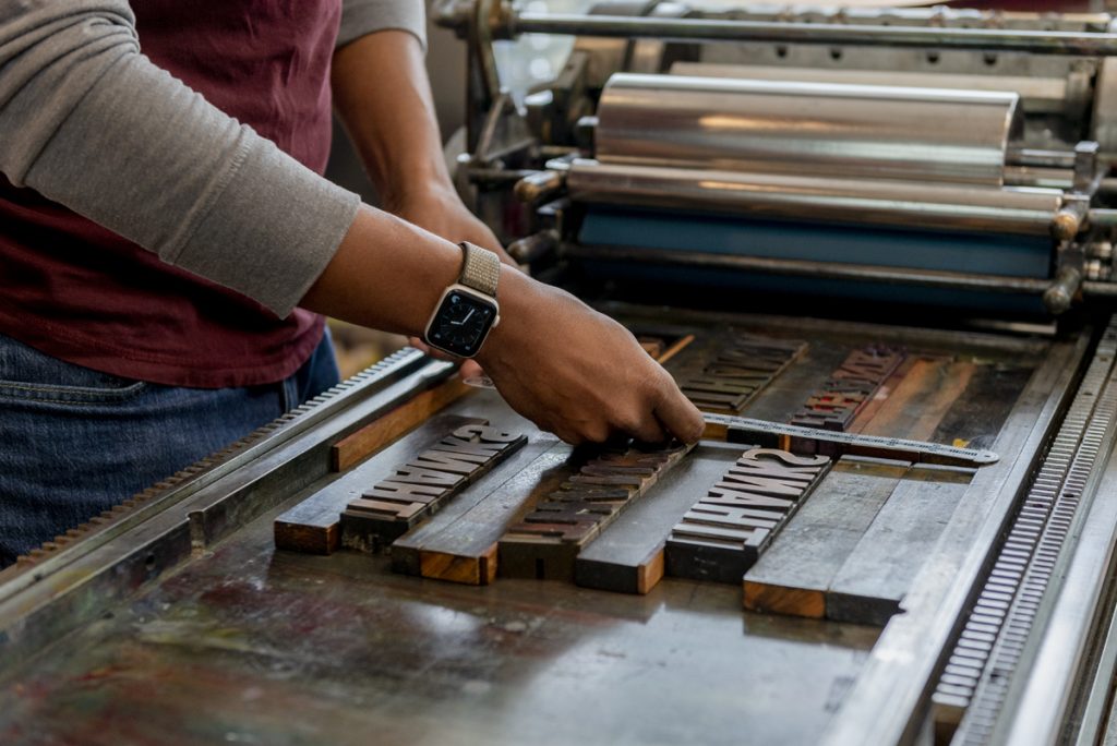 Image: Photo of artist Ben Blount's hands as he measures the spaces between letters at his letterpress. Photo by Ryan Edmund Thiel.