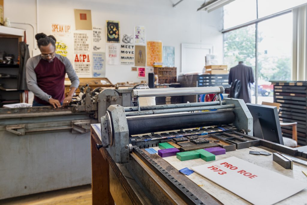 Image: Wide shot of Ben Blount working in his studio. In the foreground is a letter press with a white poster with red text that reads: "Pro Roe, Est. 1973." In the background, Ben Blount works on arranging letters in another letter press. Photo by Ryan Edmund Thiel.