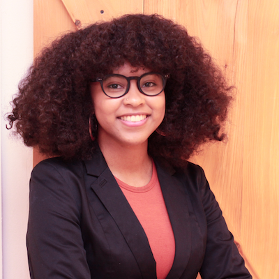 Image: A portrait of writer Chenoa Baker, who wears a dark blazer and glasses while smiling at the camera.