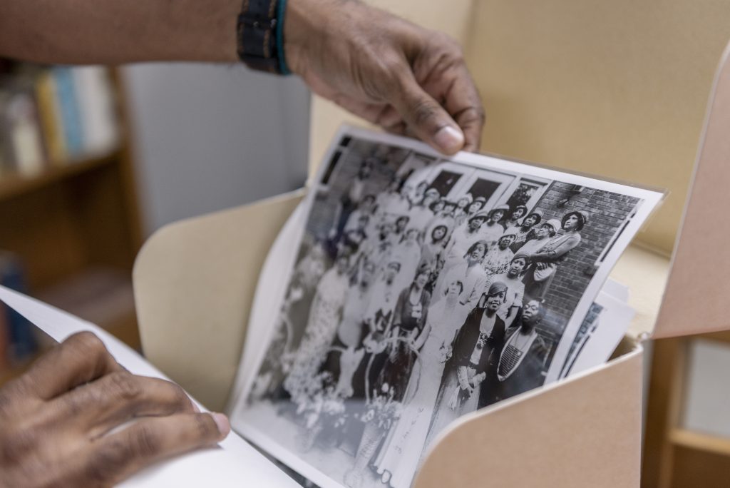 Image: Dino pulls a black and white image out of a storage container. The photograph features a large group of women posed outside of a brick building. Photo by Ryan Edmund Thiel.