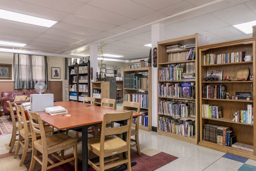 Image: The interior of Shorefront Legacy Center. A large table stands in the center of the room surrounded by seven chairs. On the table sits a large pile of papers and a white box. Lining the walls are filing cabinets and bookcases filled with books, archiving materials, boxes, photos, and memorabilia. Photo by Ryan Edmund Thiel.