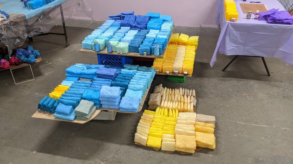 Image: Preparation of the artist's performance and art show at her studio, Fortuna called Landscape of Dual in 2023. Pallets hold blue and yellow packages organized by color. Courtesy of the artist.