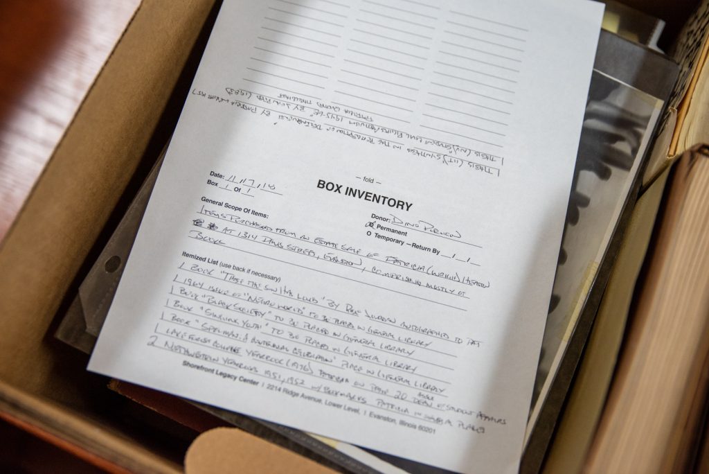 Image: A close up image inside an archival box containing photos and other papers. The top piece of paper reads: "Box Inventory" and in handwritten script below that the details of what materials can be found inside the box are written. Photo by Ryan Edmund Thiel.