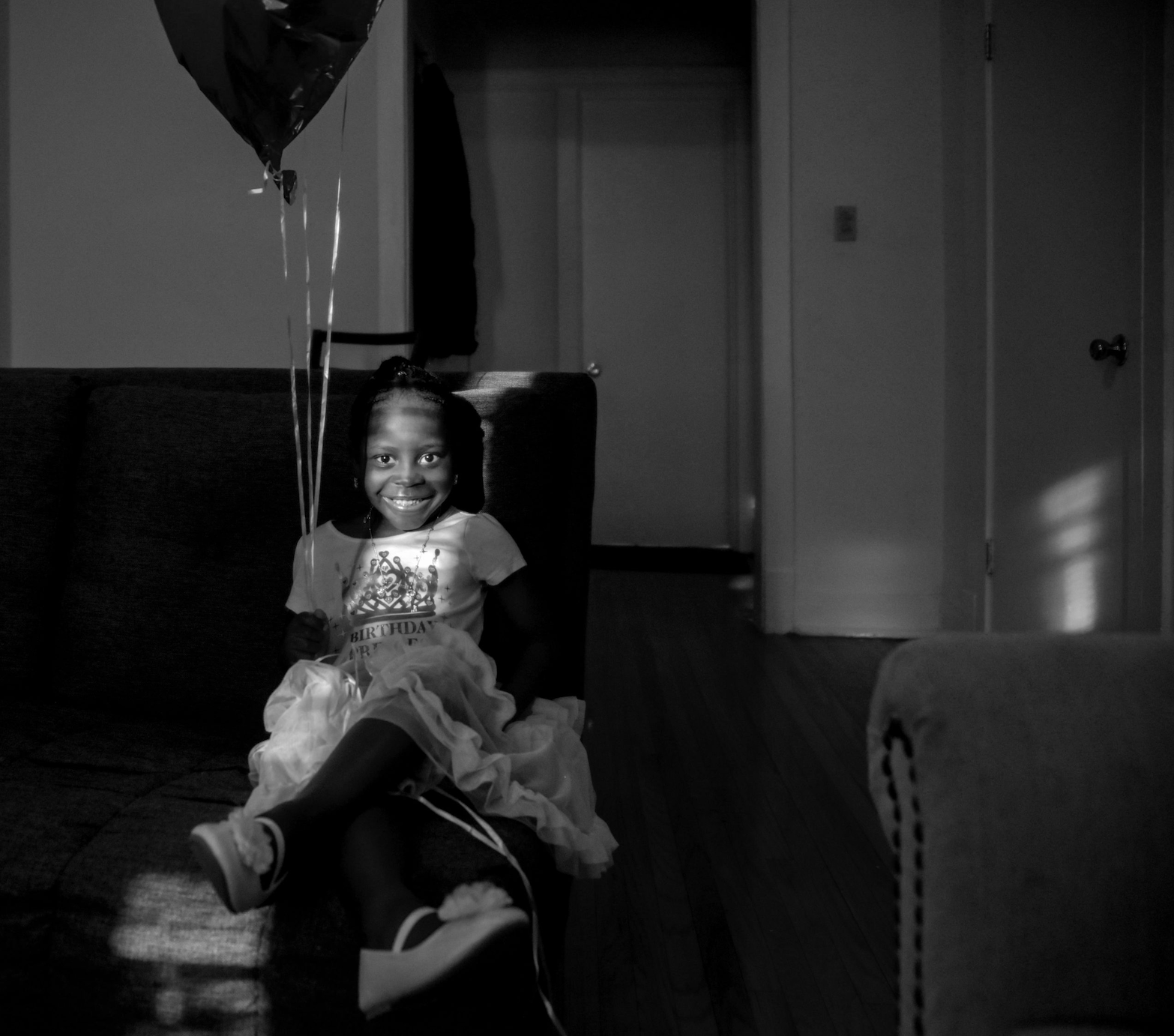 Image: birthday girl, a greyscale photograph of a young, smiling girl seated in an apartment. She holds a balloon and looks at the camera. Image by EdVetté Jones