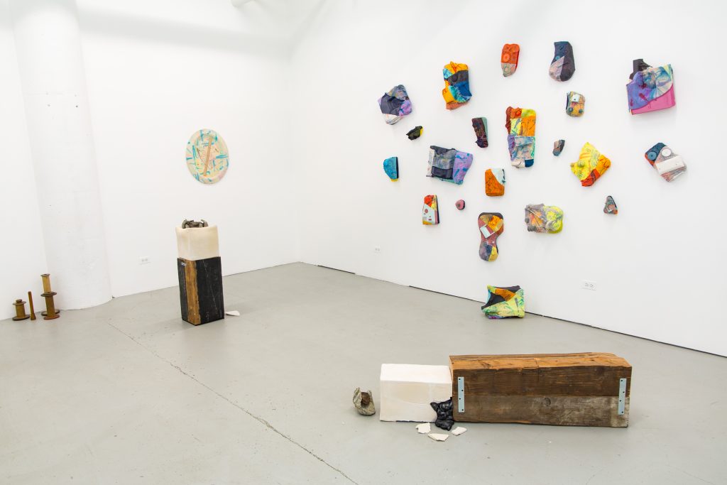 Image: Exhibition shot of For Those Who Toil (2023) at Purple Window Gallery. Both of Michael Chambers' sculptures sit left of center, while Millicent Kennedy's work frames the gallery walls. Antique thread spools are positioned on the ground near the left-hand side of the frame. Image by Amy Shelton.