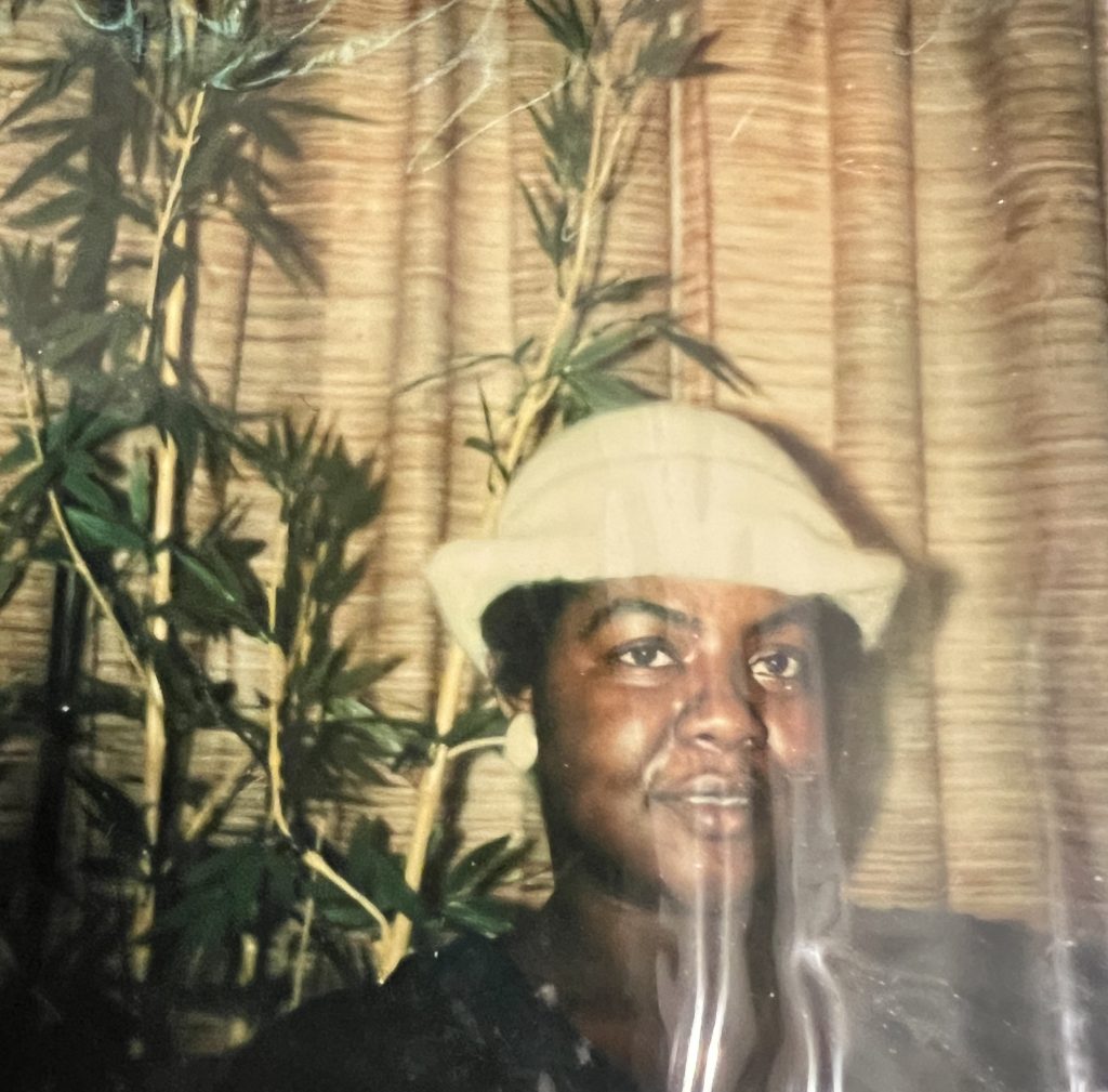 Image: Profile shot of Vanita Green. She wears a white hat and white earrings. She is framed to the left by a tall, leafy plant. Image courtesy of the Green family.
