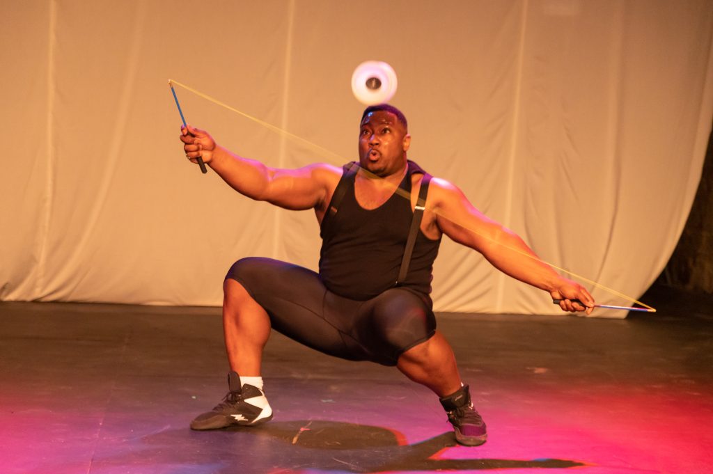 Image: A man crouches close to the ground while he has launched the diabolo in the air with a string with wands at the ends. It looks like the diabolo is balanced on his head. He is wearing a sleeveless black shirt, suspenders, and shorts. Behind him is a cream colored curtain; a pink light source lights up the floor and his body. Photo by Peter Serocki.