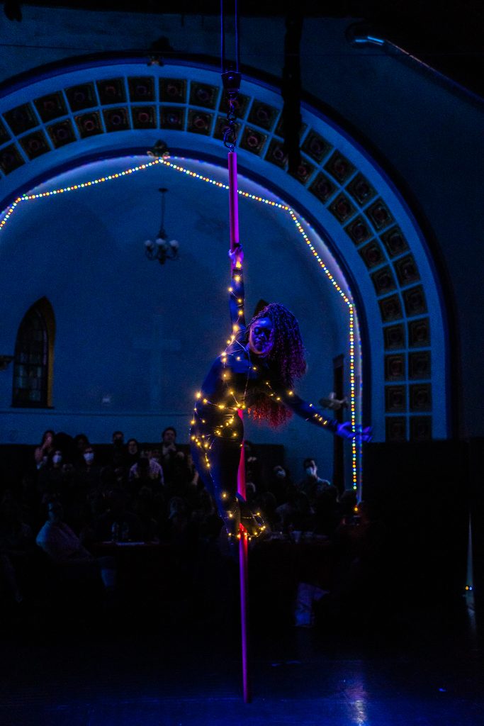 Image: A woman wrapped in golden lights poses right on a pole, holding herself up with a left arm and her feet. Behind her is an archway lit by multi-colored lights and tiles. Members of the audience can be seen in the dark behind her. Photo by Michelle Reid.