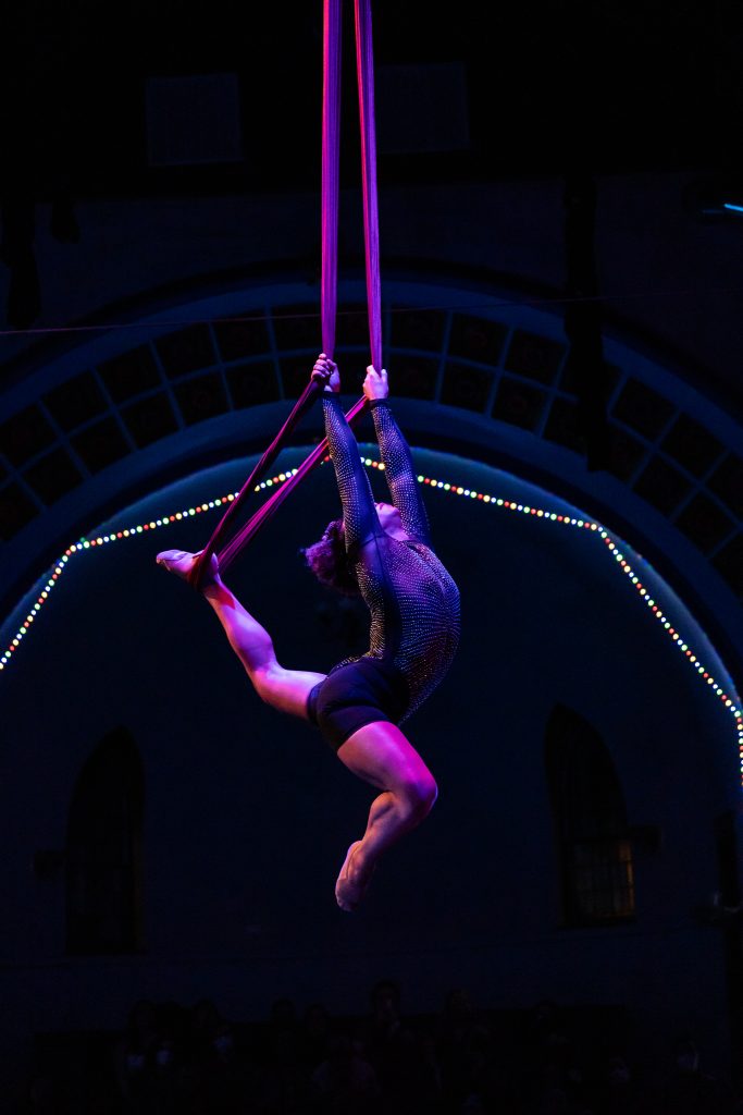 Image: Image: A person holds pink color silks with both hands while they positioned their left foot in the silks. They are arching their back, pointing their toes. They are wearing a sparkling black leotard. They are framed by an archway with multi-colored lights and tiles. The rest of the stage looks dark. Photo by Michelle Reid.