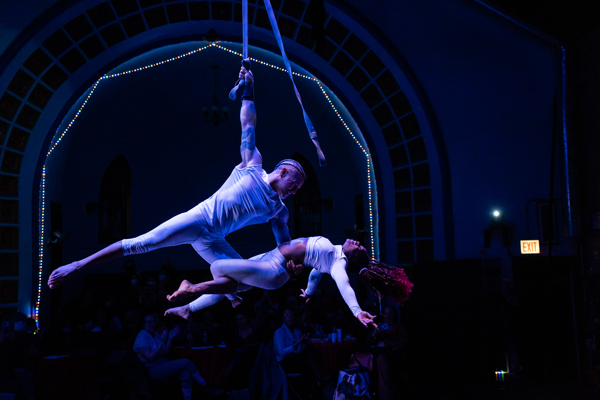 Image: A man dangles with one arm from straps, while holding a woman prone with the other arm. He has hooked one leg with hers to allow her to lean back. They are both dressed in white. They are framed by an archway with multi-colored lights and tiles. Members of the audience can be seen in the dark behind them. Photo by Michelle Reid.