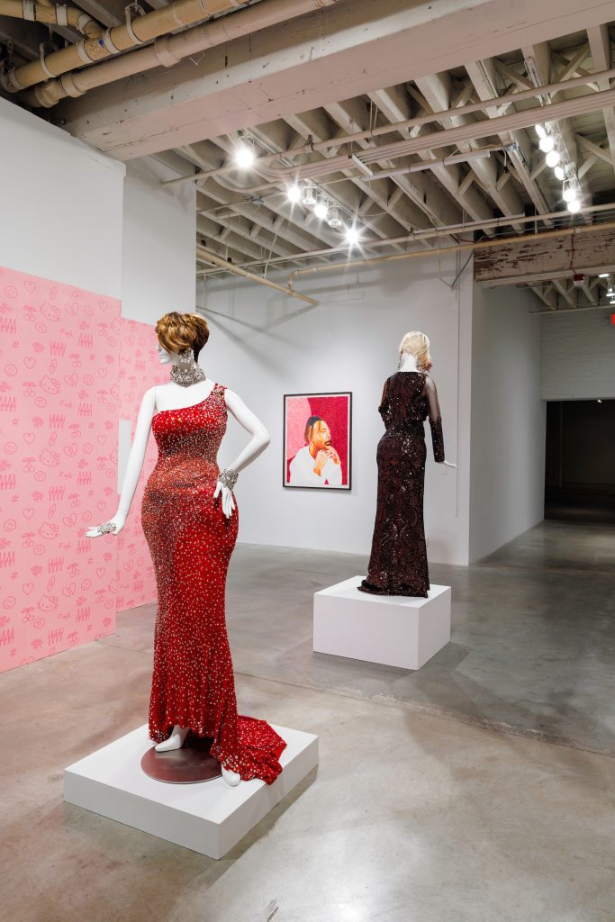 Image: Two mannequins on pedestals wearing glittering floor-length gowns. Foreground: Faleasha Savage, Expensive Addiction, 2022. Wallpaper: Yvette Mayorga, Daydreaming Notes, 2022. Background: Devan Shimoyama, Spotlight, 2022. Image courtesy of Bemis Center.