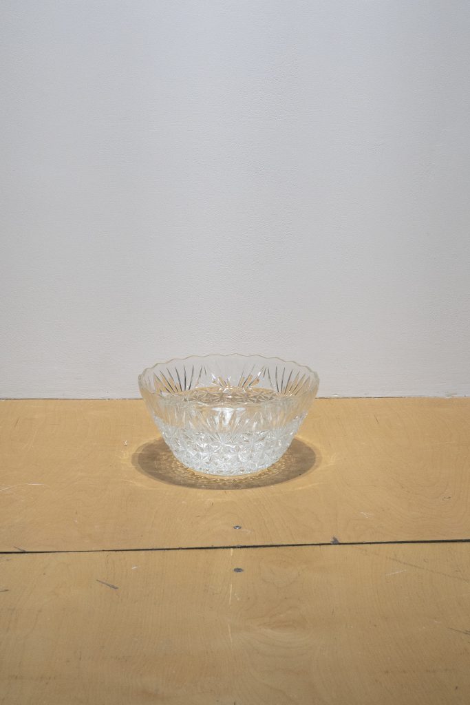 Image: Still shot of Devin T. Mays, At rest, a vessel, 2022. Vessels, water. Courtesy the artist. Abridged text of verbal description of the artwork courtesy of Gallery400: three water vessels, one made of clear crystal glass in the shape of a round bowl that is 6 inches high; one made of white plastic in the shape of a rectangle that is 5¾ inches high; and one made of green plastic in the shape of a round bowl that is 3¾ inches high, are placed in different areas of the gallery’s floor. The glass vessel is placed against the back wall of the main gallery room, the white vessel is placed against the north wall in the main gallery room, and the green vessel is placed next to a cylindrical white column. Water levels inside these containers fall during the exhibition depending on evaporation rates, and are refilled by gallery staff members, according to the artist’s instruction.