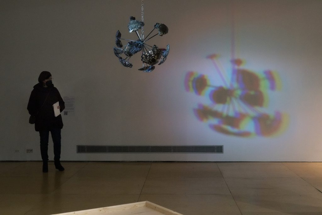 Image: Young Joon Kwak, Brown Rainbow Eclipse Explosion, 2017. Cast aluminum, welded aluminum rods, resin, glass mirror tiles, acrylic mirrors, epoxy putty, acrylic paint, motor, steel chain and hardware, custom LED light, shadow. Courtesy the artist and Commonwealth and Council, Los Angeles and Mexico City.
Abridged text of verbal description of the artwork courtesy of Galllery400: a large, silver disco ball that has exploded is suspended by a metal chain on the ceiling. It hangs approximately six feet high from the floor. On the disco ball’s surface are mosaic pieces of a mirror reflecting light. The fragmented pieces of the disco ball expose its rough textured insides and are held together by individual aluminum  rods welded together at the center. The thick aluminum rods vary in their length, some are long while others are short.
The disco ball slowly spins and is illuminated by a multicolored light source secured on the ceiling. On the wall, is the disco ball’s enlarged shadow.