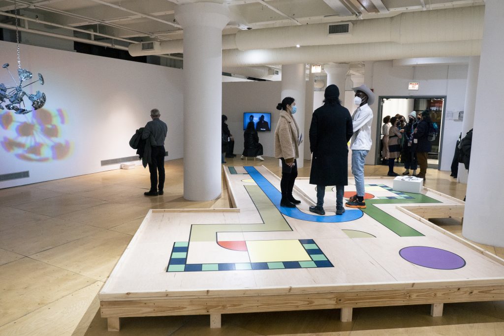 Image: Exhibition shot of Reckless Rolodex, courtesy of Gallery400. 3 visitors stand atop FAKE's platform, while Young Joon Kwak's work hangs to the left in the background. Joon Kwak's piece projects a shadow on the gallery wall. Right of center, a screen plays Yu's video piece. Gallery visitors linger in hallway that leads to antechambers filled with Steger's archives and the work of other artists.