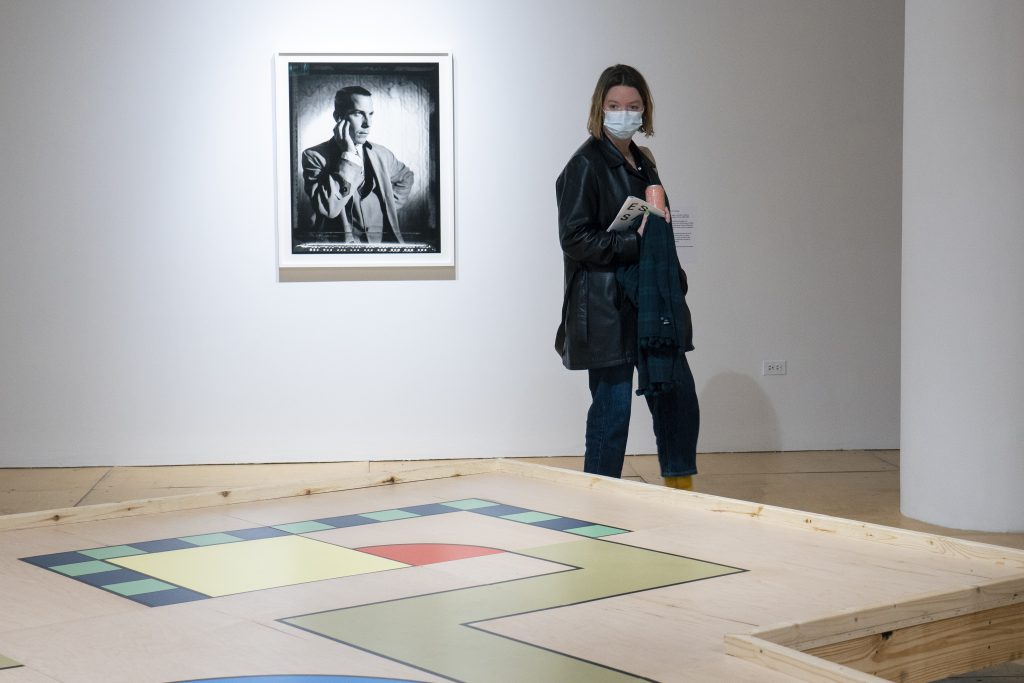 Image: Exhibition shot of Reckless Rolodex, courtesy of Gallery400.  A portrait of Lawrence Steger hangs alone, centered, on gallery wall, as a visitor looks down at FAKE's platform. 