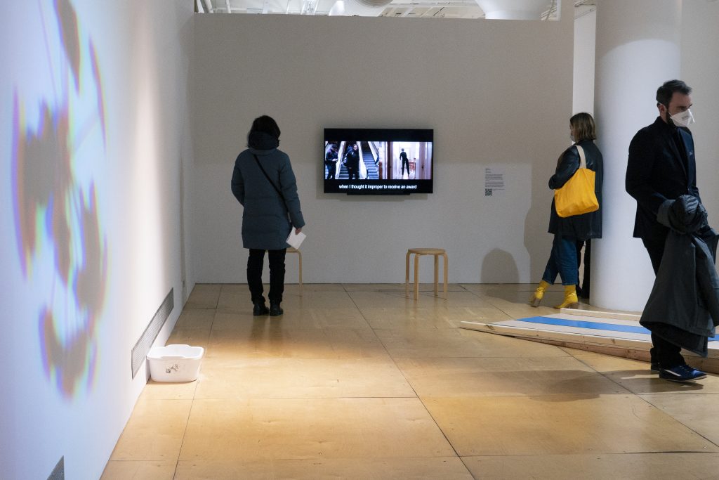 Image: Exhibition image of Cherrie Yu, Trisha and Homer 2018. Video, 9:05 mins. Courtesy the artist.
Abridged text of verbal description of the artwork courtesy of Gallery400: color film begins in a two-channel format featuring two women in either frame cropped at the waist. Found footage of the dancer-choreographer Trisha Brown’s 1986 performance, Accumulation with Talking Plus Watermotor, plays on the left. In this frame the artist, Trisha Brown performs a series of movements in a dance studio, speaking slowly. On the right, is the contemporary artist Cherrie Yu. Yu replicates Brown’s dance and the two figures produce the same series of movements simultaneously. Brown’s voice alone is audible, even after the video switches to a single-channel format. In the next scene, the video resumes a two-channel structure. Brown’s voice remains audible. The left-hand frame features a public stairway running between two escalators (one up, one down). As people pass up and down the passageways, the camera remains centered on the back of Homero Muñoz mopping the stairs with a mop. Muñoz has short brown hair, wears navy pants, a loose navy jacket, sneakers, and appears to be at work. In the right-hand screen, the camera centers on the back of Yu in a kitchen. Yu wears the same black shirt and pants from the earlier sequence. She has no mop but reproduces the actions of the Muñoz in time with his own. The final portion of the film depicts Yu and Muñoz seated against a wall. They face the camera without speaking and move very little. Yu, on the left, wears a white shirt over a black tank top and glasses. Muñoz wears a black button up shirt with a white undershirt. They appear to be watching the same film,Trisha and Homer. Although Yu and Muñoz do not speak in the frame, subtitles capture their audible discussion about dance, labor, music, and performance. 