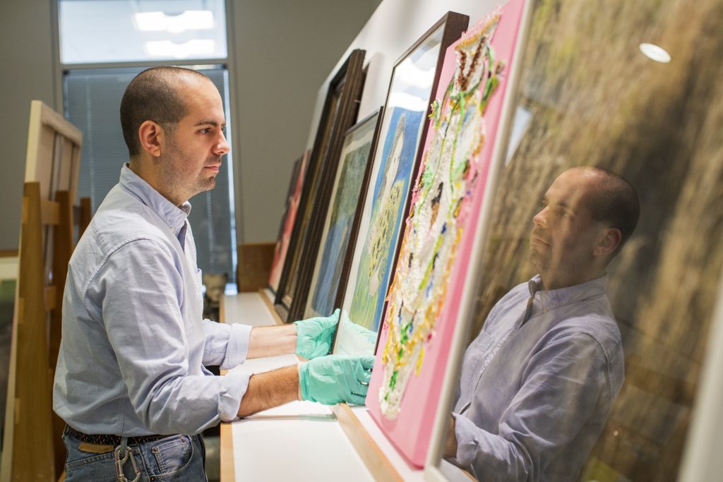 Image: David Maruzzella stands in front of a series of artworks. He is wearing a light blue shirt, jeans, and blue-green gloves as he picks up one of the pieces. Photo by Kristie Kahns.