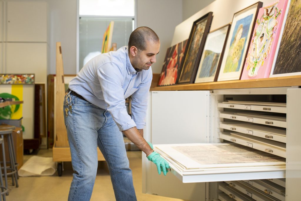 Image: David Maruzzella bends over a pull-out drawer in the DePaul Art Museum Archives. He is wearing a light blue shirt, jeans, and blue-green gloves as he opens one of the drawers. Photo by Kristie Kahns.