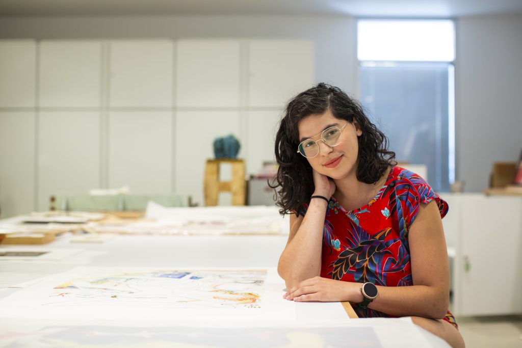 Image: Natasha Mijares resting her elbow on a table and holding her neck. She looks at the camera, smiling gently. Photograph by Kristie Kahns.