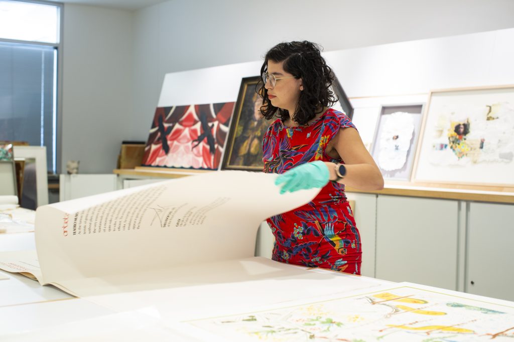 Image: Natasha Mijares turning a page of a large pad, obscuring the page she is looking at. There is text on the page facing us. Photograph by Kristie Kahns.