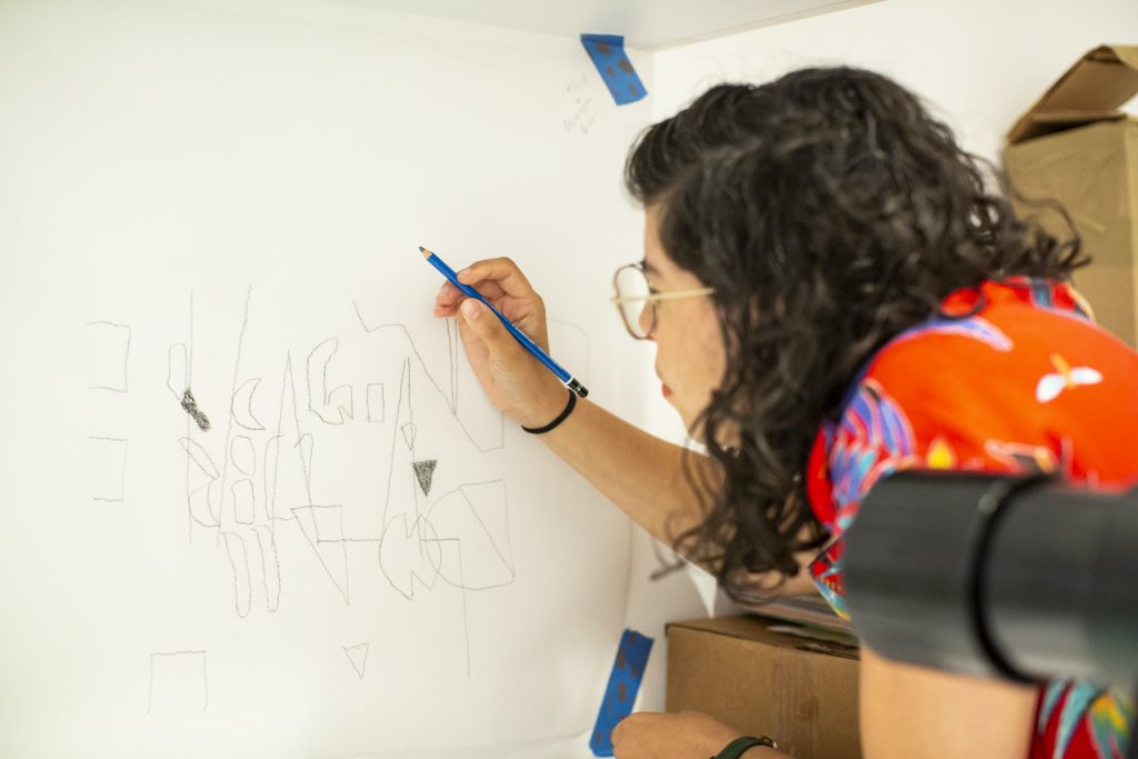 Image: Natasha hunches towards a wall holding a pencil in her right hand. Blue electrical tape holds a piece of paper with scribbled shapes in the center of it. Photograph by Kristie Kahns.