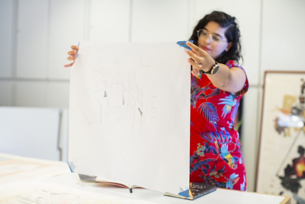 Image: Natasha Mijares stands and holds a square piece of paper in front of her by the corners. The paper has scribbled lines and shapes on it. Photograph by Kristie Kahns.