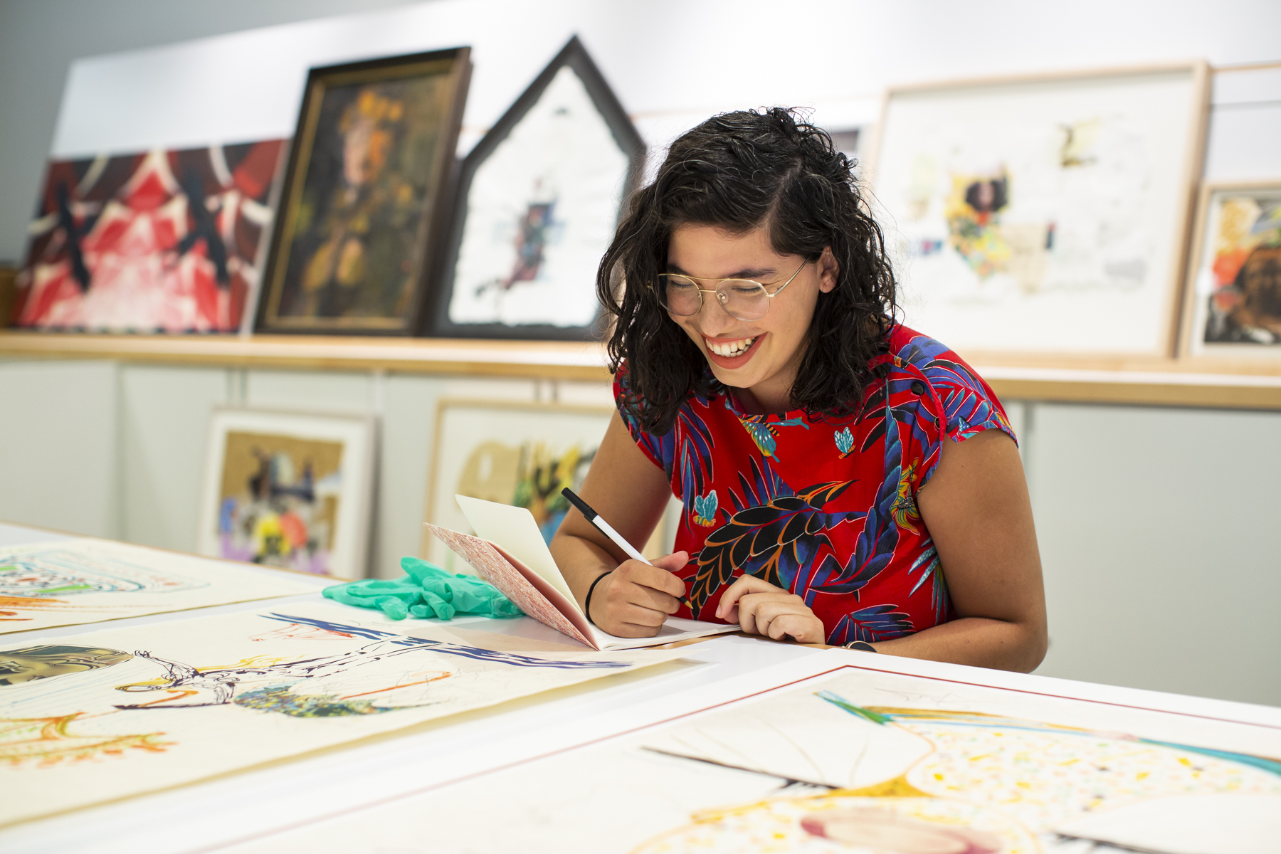 Image: Natasha Mijares smiling and seated at a desk, leaning gently forward. She holds a pen and notebook in her right hand. On the desk are prints and other art works. Behind Natasha are shelves holding artworks. Photograph by Kristie Kahns.