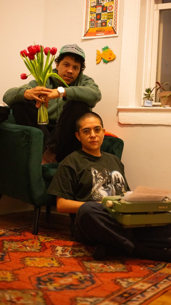 Image: Ezra Amiri and Remy Guzman in Stuti Sharma's living room. Ezra is wearing a green hooded sweatshirt and sitting on a green chair holding a bouquet of lilies, Remy is wearing a shirt which depicts a wolf, the moon, and trees, they are sitting in front of the chair holding a green typewriter on their lap. There is a patterned orange rug covering most of the floor which they are both on. There is a poster a cut out fish and two potted plants on the left side of Remy and Ezra. Photo by Stuti Sharma.