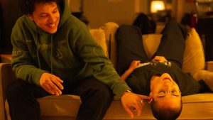 Image: A photograph of two people, (the directors of Lost & Found), sitting on a light brown couch. The person on the left, Ezra Amiri, is wearing a green sweatshirt and has curly hair. He is sticking his finger in Remy Guzman's ear playfully. Remy has a buzzcut and is sitting upside down on the couch.