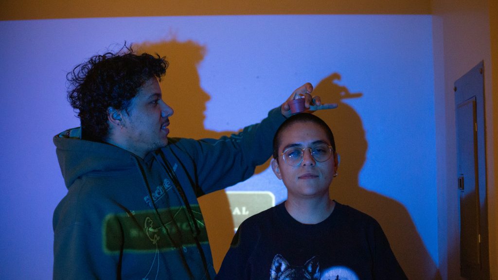 Image: Ezra Amiri and Remy Guzman pictured in Stuti Sharma''s apartment. Ezra is wearing a green hoodie and placing a small potted plant on Remy's head. Remy is wearing a black shirt which has a wolf, trees and a moon. They are standing in front of a blue projector offline screen. Photo by Stuti Sharma.