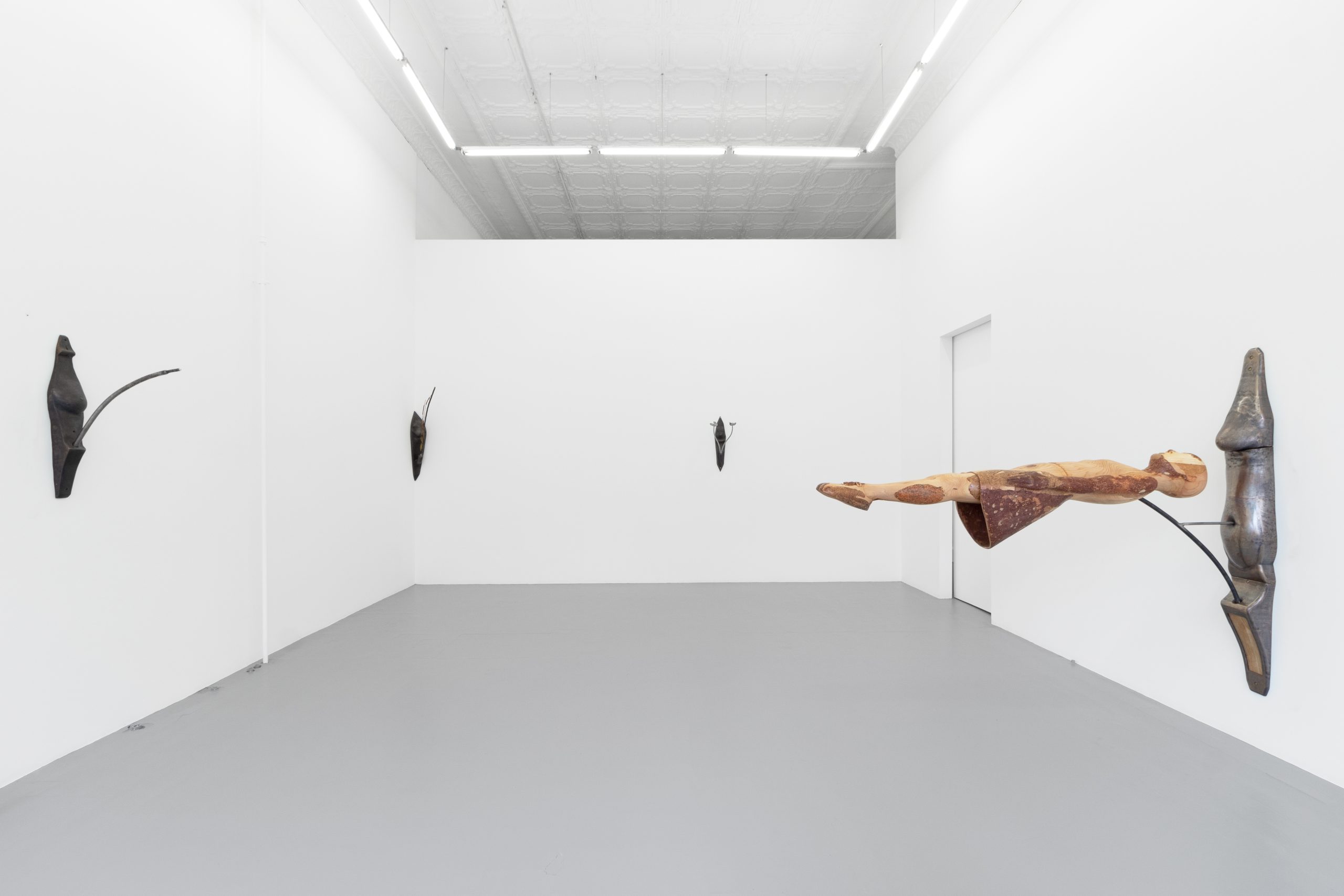 Image: Installation view of Basket Toss. A girl made from bronze and wood floats horizontally, affixed to the righthand wall. Metal breasts dot the other two walls, intertwined with mushrooms and cattails.