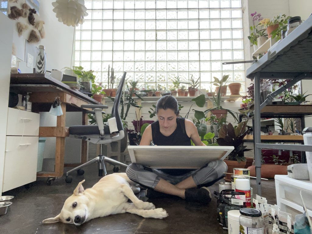 Image: Self portrait of Laleh Motlagh in her studio. She is surrounded by plants, art supplies, and her dog. Image courtesy of Laleh Motlagh.