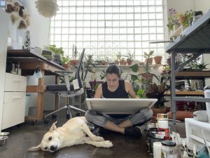 Image: Laleh Motlagh's studio. In the background, a gridded glass window. Around it are houseplants. Seated on the concrete floor in the foreground is Motlagh, with a board on her crossed legs. Laying on their to side to Motlagh's side is a large white dog. Photo courtesy of the artist.
