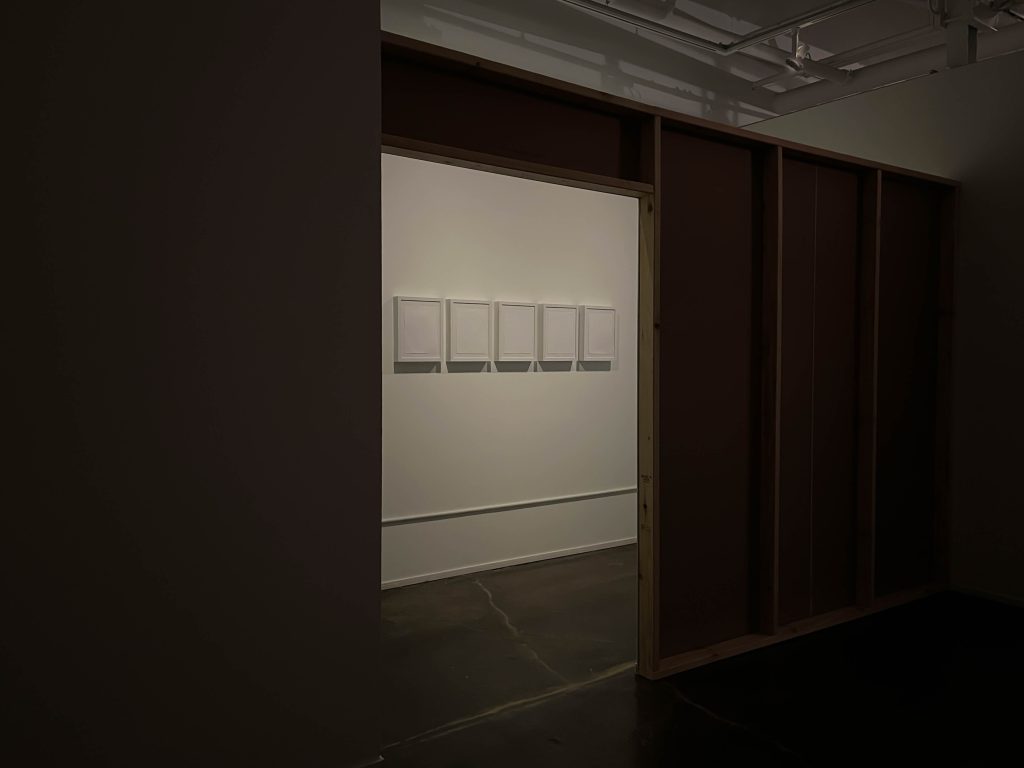 Image: An installation photo of Contemplation. There is an entryway made of wood in the foreground. Through the entryway, we can see five sheets of paper framed and hung. Image courtesy of Laleh Motlagh.